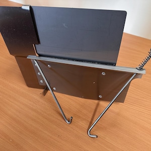 May include: A metal fire pit wind guard with a black rectangular shield and a silver-colored frame. The wind guard has two angled support legs and a spring-loaded adjustment mechanism.