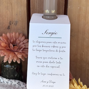 Puede incluir: Un tapón de botella con un diseño decorativo, sujeto por una tarjeta blanca con el nombre "Sergio" y texto en español. La tarjeta se coloca frente a un fondo de madera, con flores en primer plano.