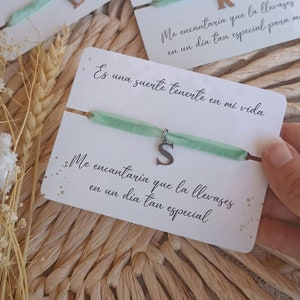 Puede incluir: Una tarjeta blanca con el texto "Me encantaria que la llevases en un dia tan especial para mi" y "Es una suerte tenerte en mi vida". Una cinta verde con un dije de plata con la letra "S" está unida a la tarjeta.