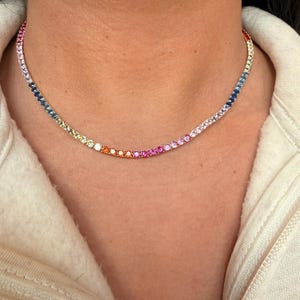 May include: A vibrant tennis necklace displaying a rainbow gradient of gemstones. The necklace showcases a spectrum of colours, including pink, orange, yellow, green, blue, and purple. The necklace is worn around the neck.