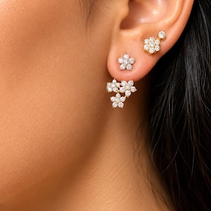 Puede incluir: Un conjunto de tres pendientes dorados con diseños en forma de flor incrustados con piedras transparentes y brillantes. Incluye un pendiente pequeño, uno de flor más grande y un pendiente con diseño floral que cuelga bajo el lóbulo.