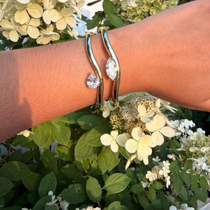May include: Two gold-toned metal cuff bracelets with a wavy, curved design. One bracelet features a single pear-shaped clear crystal, while the other bracelet features a single marquise-shaped clear crystal, both set on the ends of the metal bands.