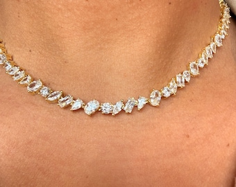 May include: A gold-toned necklace featuring a continuous row of clear, multi-shaped faceted stones. The stones include marquise, oval, pear, and rectangular cuts, all set in a linked metal chain that rests closely around the base of the neck.