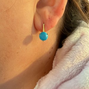 Puede incluir: Un pendiente de color dorado con una piedra turquesa brillante. El pendiente es de estilo palanca, con la piedra turquesa sujeta por pequeñas garras. El pendiente se muestra en la oreja de una persona.