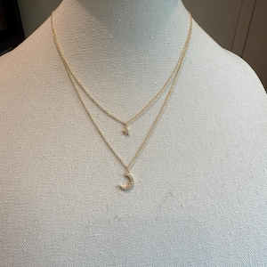 Op de afbeelding: Een goudkleurige, gelaagde ketting met een kleine sterhanger en een halve maan hanger. De ketting wordt getoond op een witte mannequin. De maan hanger is bezet met kleine, heldere stenen.