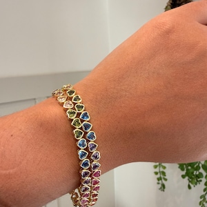 May include: Gold-toned bracelet featuring heart-shaped gemstones in a rainbow of colors. The bracelet is composed of multiple rows of the heart-shaped stones, creating a vibrant and eye-catching accessory. The gemstones include shades of pink, purple, blue, green, and yellow.