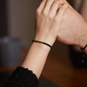 May include: Two people holding hands, each wearing a black beaded bracelet with a black cord.