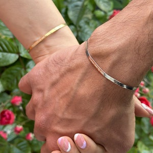 Puede incluir: Dos personas se toman de la mano, cada una lleva una fina pulsera de plata con una inscripción. La pulsera de la mano izquierda dice "Siempre lado a lado". La persona de la derecha lleva una pulsera de oro en la muñeca.