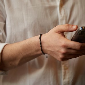 Könnte beinhalten: Ein schwarzes Perlenarmband mit einer Schnurverschlusses. Das Armband wird am Handgelenk einer Person getragen, die ein weißes Hemd trägt.