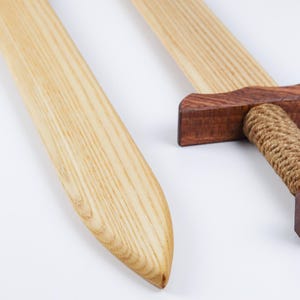 May include: Two wooden swords featuring light-colored blades with visible grain patterns. Each sword has a dark brown wooden crossguard and a handle wrapped in thick, tan-colored rope. The blades are smooth and tapered toward the rounded points.