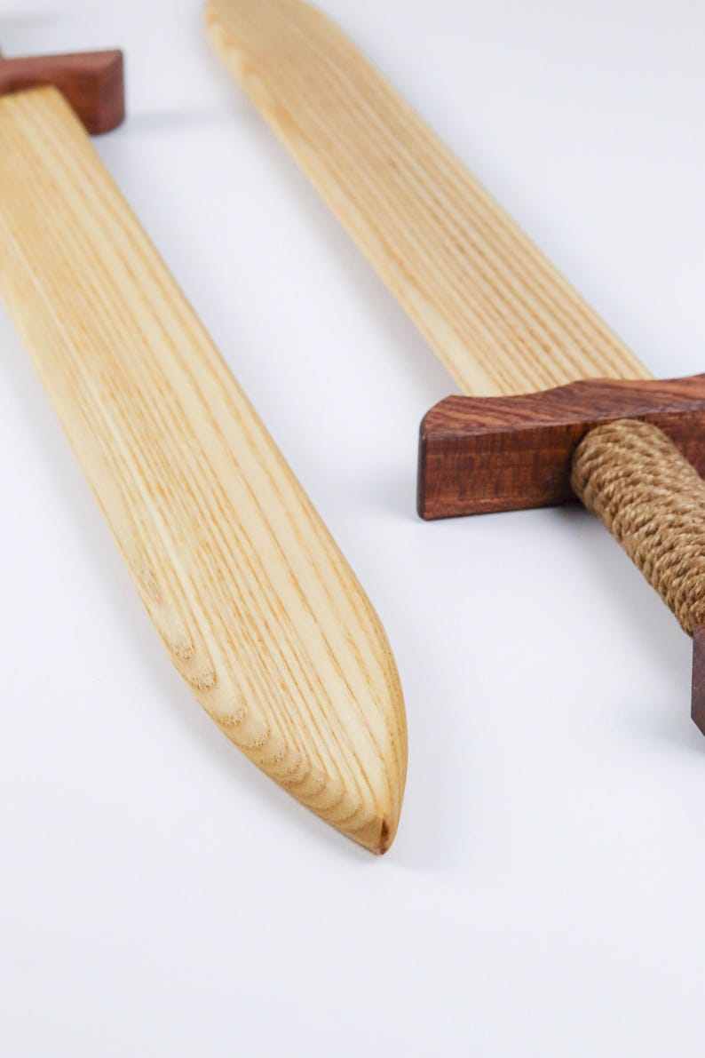 May include: Two wooden swords featuring light-colored blades with visible grain patterns. Each sword has a dark brown wooden crossguard and a handle wrapped in thick, tan-colored rope. The blades are smooth and tapered toward the rounded points.