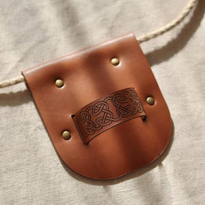 May include: A small, brown leather pouch with a decorative Celtic knot design on a band. The pouch has a rounded bottom and is secured with brass rivets. A rope is attached to the top.