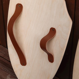 May include: A light-coloured wooden shield with a pointed bottom edge and a curved top. The shield features two brown leather straps attached to the front surface with small metallic rivets, providing a grip for holding the wooden object securely.