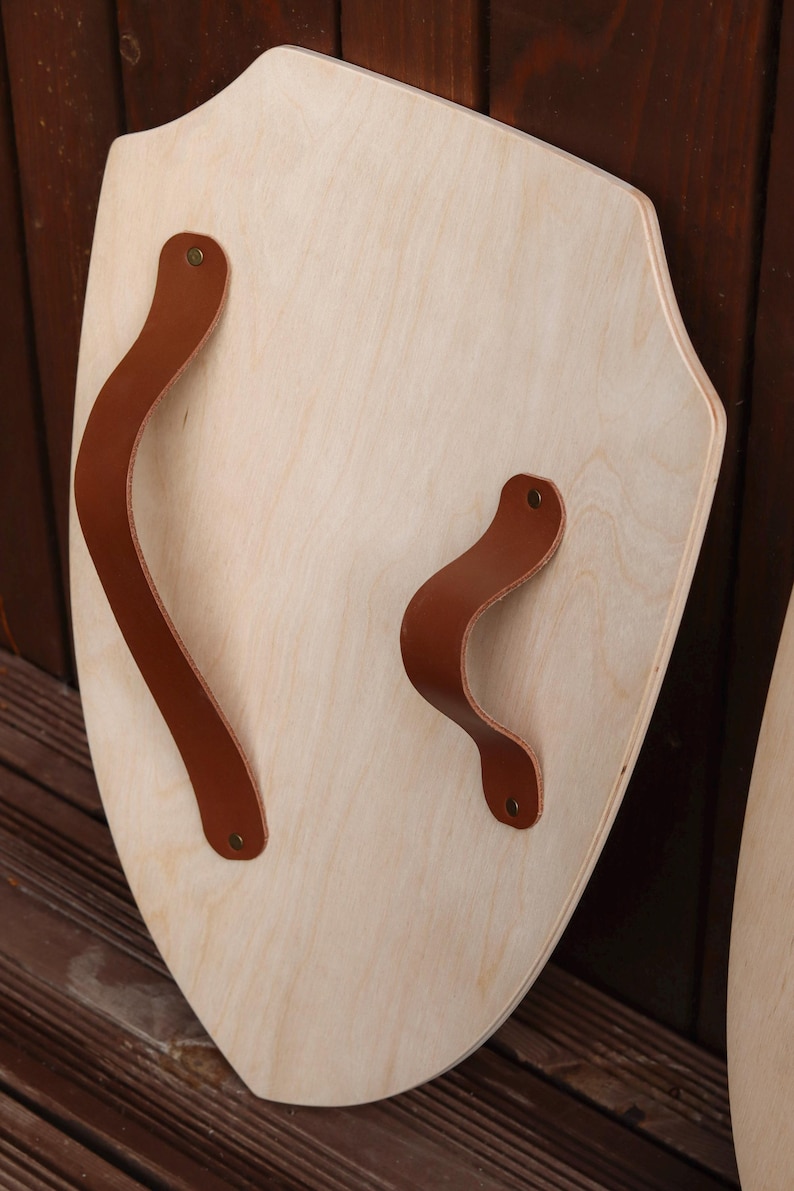 May include: A light-coloured wooden shield with a pointed bottom edge and a curved top. The shield features two brown leather straps attached to the front surface with small metallic rivets, providing a grip for holding the wooden object securely.