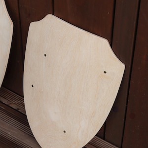 May include: A light-coloured wooden shield with a smooth, unfinished surface and a pointed bottom edge. The shield features three small, evenly spaced circular holes drilled through the wood, arranged in a triangular pattern across the centre.