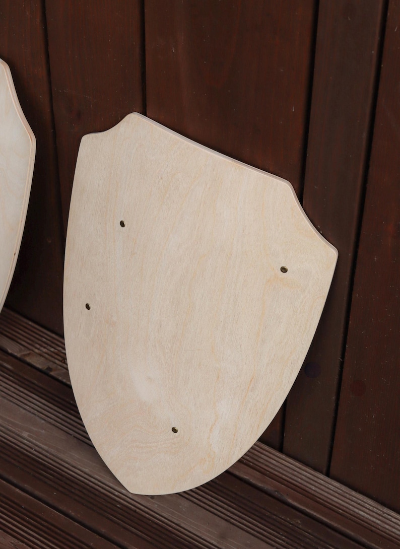 May include: A light-coloured wooden shield with a smooth, unfinished surface and a pointed bottom edge. The shield features three small, evenly spaced circular holes drilled through the wood, arranged in a triangular pattern across the centre.