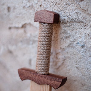 May include: A wooden toy sword featuring a dark brown polished wood pommel and crossguard. The handle is wrapped in thick, light tan natural fiber rope. The blade is made of a lighter-toned, smooth wood with a visible straight grain pattern throughout.