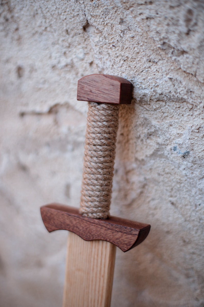 May include: A wooden toy sword featuring a dark brown polished wood pommel and crossguard. The handle is wrapped in thick, light tan natural fiber rope. The blade is made of a lighter-toned, smooth wood with a visible straight grain pattern throughout.