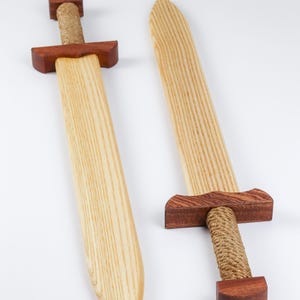 May include: Two wooden swords featuring light-colored blades with visible wood grain, dark reddish-brown wooden crossguards and pommels, and handles wrapped in thick, natural-colored jute rope. The swords are positioned side by side.
