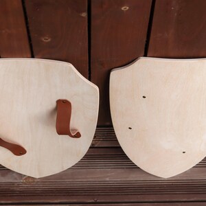 May include: Two light-coloured wooden shields with a smooth finish. One shield features two brown leather straps attached with metal rivets for holding, while the other shield shows four small metal rivets visible on the front surface.