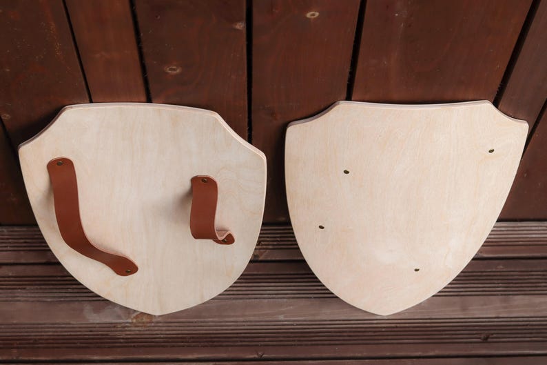 May include: Two light-coloured wooden shields with a smooth finish. One shield features two brown leather straps attached with metal rivets for holding, while the other shield shows four small metal rivets visible on the front surface.