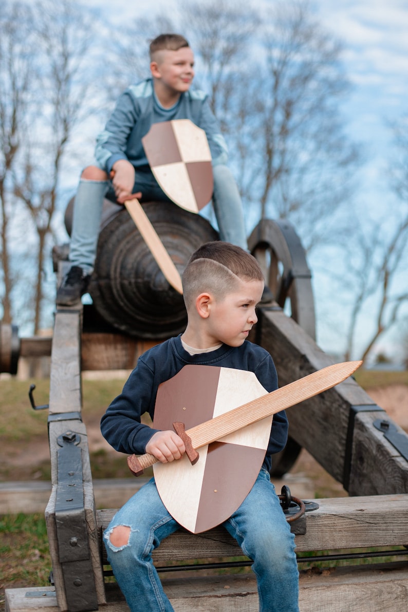 May include: A wooden toy sword and a shield with a quartered brown and light wood pattern. The sword features a straight blade and a crossguard, while the shield has a smooth, flat surface. These items are designed for imaginative play.