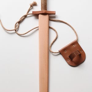 May include: A wooden toy sword with a light-colored blade and a darker wood pommel. The handle is wrapped in thick tan rope. A matching tan rope connects the sword to a brown leather belt holster featuring a small embossed knotwork pattern.