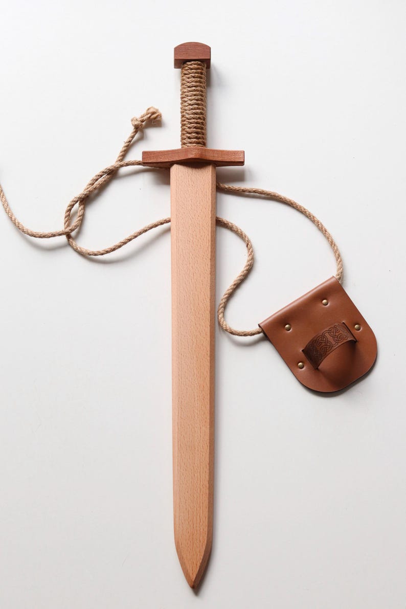 May include: A wooden toy sword with a light-colored blade and a darker wood pommel. The handle is wrapped in thick tan rope. A matching tan rope connects the sword to a brown leather belt holster featuring a small embossed knotwork pattern.