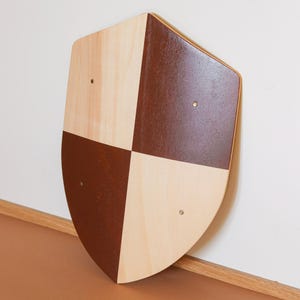 May include: A shield-shaped wooden plaque featuring a quartered design with alternating light natural wood grain and solid dark brown painted sections. Four small metallic rivets are positioned at the center of each quadrant on the surface.