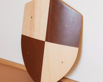 May include: A shield-shaped wooden plaque featuring a quartered design with alternating light natural wood grain and solid dark brown painted sections. Four small metallic rivets are positioned at the centre of each quadrant on the surface.