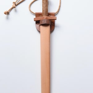 May include: A wooden toy sword with a light brown finish, featuring a handle wrapped in tan rope. The sword is held in a tan leather scabbard with a dark brown leather crossguard detail embossed with a Celtic knot pattern and a tan rope neck strap.
