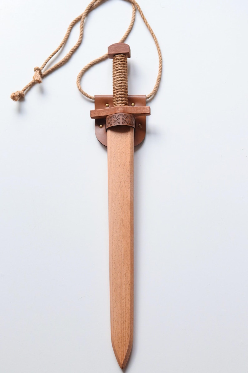 May include: A wooden toy sword with a light brown finish, featuring a handle wrapped in tan rope. The sword is held in a tan leather scabbard with a dark brown leather crossguard detail embossed with a Celtic knot pattern and a tan rope neck strap.