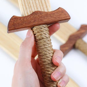 May include: A wooden toy sword featuring a light-colored blade with a visible wood grain, a dark brown crossguard, a handle wrapped in thick tan rope, and a matching dark brown pommel at the base of the hilt, with additional swords visible.