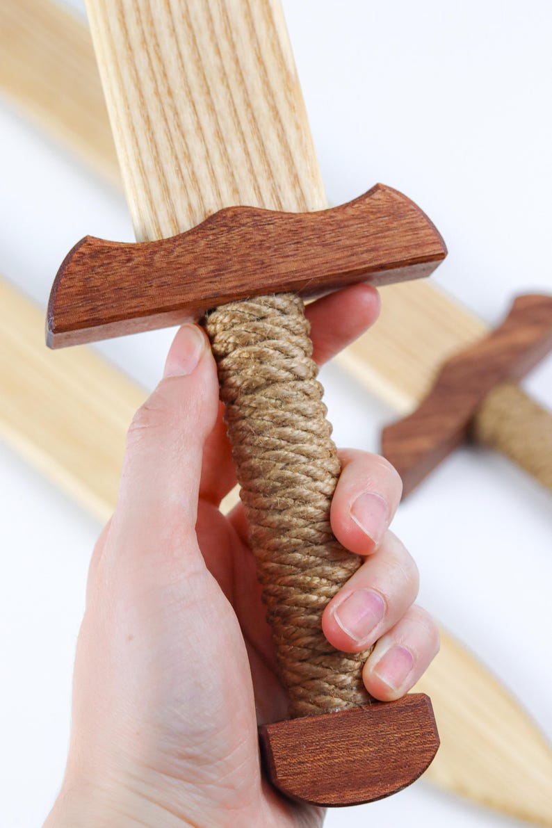 May include: A wooden toy sword featuring a light-colored blade with a visible wood grain, a dark brown crossguard, a handle wrapped in thick tan rope, and a matching dark brown pommel at the base of the hilt, with additional swords visible.