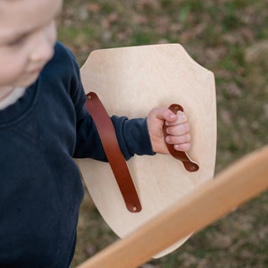 May include: A light-coloured wooden shield with a smooth finish, featuring a brown leather strap handle secured by metal rivets. The shield has a classic pointed base and a curved top edge, designed for holding with a single hand.