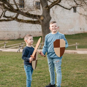 May include: A wooden toy sword and a wooden toy shield with a brown and tan checkered pattern. The sword has a long, flat blade and a cross-guard handle. The shield is shaped like a heater shield with a flat top and a pointed bottom edge.