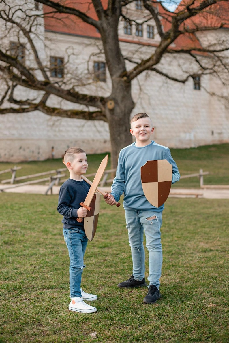 May include: A wooden toy sword and a wooden toy shield with a brown and tan checkered pattern. The sword has a long, flat blade and a cross-guard handle. The shield is shaped like a heater shield with a flat top and a pointed bottom edge.