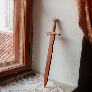 May include: A wooden toy sword with a smooth, reddish-brown blade and a crossguard featuring a central metal rivet. The handle is wrapped in thick, light-coloured natural fibre rope, and the pommel is a simple, rectangular wooden block at the top.