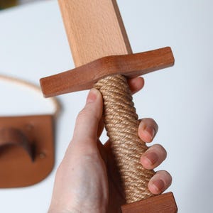 May include: A wooden toy sword featuring a light-colored blade, a darker brown wooden crossguard, and a handle wrapped in thick, tan-colored rope. The pommel at the base of the handle is made of matching dark brown wood, providing a sturdy grip.