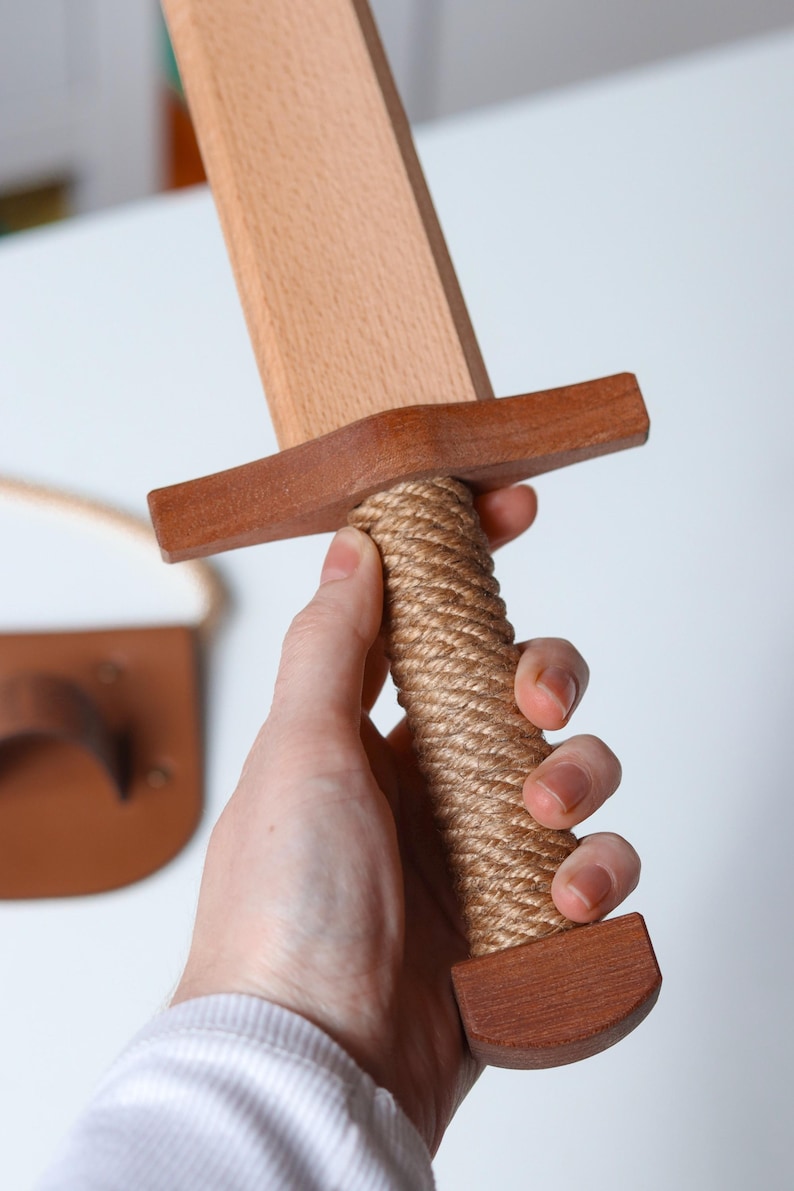 May include: A wooden toy sword featuring a light-colored blade, a darker brown wooden crossguard, and a handle wrapped in thick, tan-colored rope. The pommel at the base of the handle is made of matching dark brown wood, providing a sturdy grip.