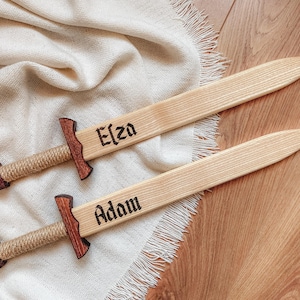 May include: Two wooden toy swords with light-colored blades and dark-stained crossguards. The handles are wrapped in tan rope. The blades are engraved with the names Elza and Adam in a black gothic-style font, featuring pointed tips.