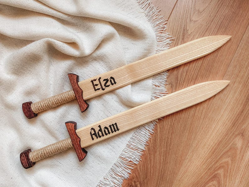 May include: Two wooden toy swords with light-colored blades and dark-stained crossguards. The handles are wrapped in tan rope. The blades are engraved with the names Elza and Adam in a black gothic-style font, featuring pointed tips.