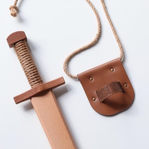 May include: A wooden toy sword with a rope-wrapped handle and a matching brown leather scabbard featuring an embossed Celtic knot pattern. The scabbard is attached to a long, natural-colored rope cord for carrying, providing a complete set.