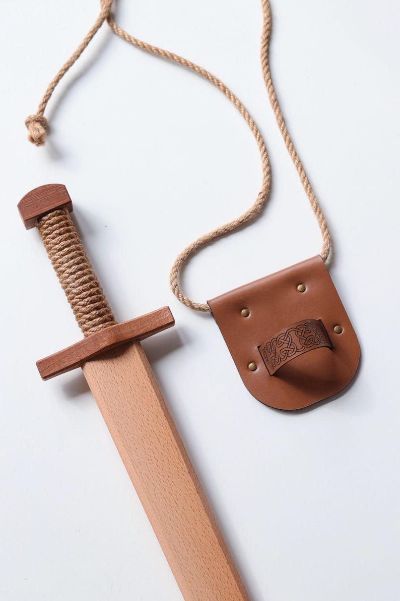 May include: A wooden toy sword with a rope-wrapped handle and a matching brown leather scabbard featuring an embossed Celtic knot pattern. The scabbard is attached to a long, natural-colored rope cord for carrying, providing a complete set.