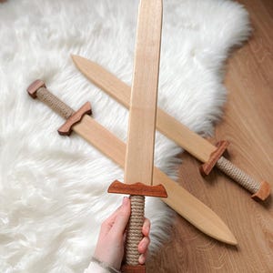 May include: Two wooden toy swords featuring light-colored blades and darker wood crossguards. The handles are wrapped in thick, light-tan rope, and the pommels are made of matching dark wood. The blades have a smooth, tapered, pointed design.