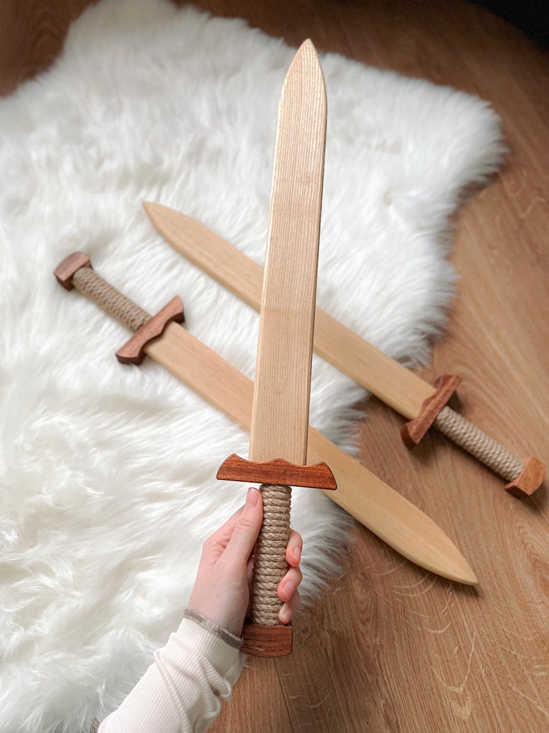 May include: Two wooden toy swords featuring light-colored blades and darker wood crossguards. The handles are wrapped in thick, light-tan rope, and the pommels are made of matching dark wood. The blades have a smooth, tapered, pointed design.