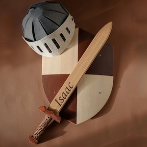 May include: A grey felt knight helmet, a wooden shield with a brown and cream quartered pattern, and a wooden sword with a rope-wrapped handle. The sword is engraved with the name Isaac in a dark script font, featuring a dark wood crossguard.