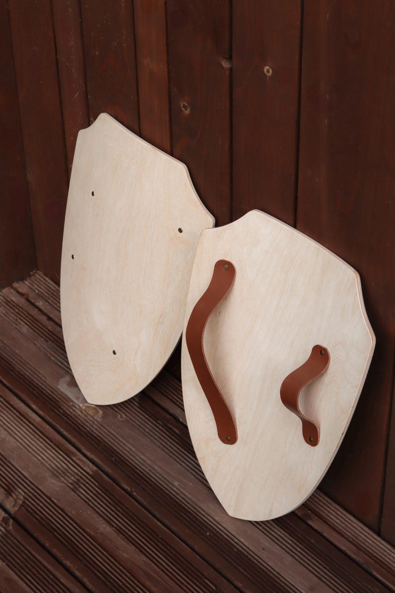 May include: Two light-coloured wooden shields with a smooth finish and a pointed bottom edge. One shield features a brown leather handle attached with metal rivets, while the other shield displays three small circular holes on its surface.