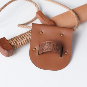 May include: A tan leather belt frog featuring a central loop embossed with a dark brown Celtic knot pattern, secured by four brass-colored metal rivets. A wooden handle wrapped in thick tan rope is positioned behind the leather accessory.
