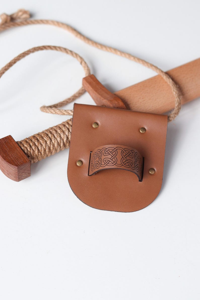 May include: A tan leather belt frog featuring a central loop embossed with a dark brown Celtic knot pattern, secured by four brass-colored metal rivets. A wooden handle wrapped in thick tan rope is positioned behind the leather accessory.
